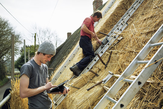 Two thatchers thatching a roof, one using a digital tablet the other standing on a ladder.