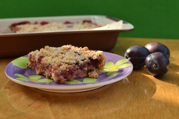 Piece of warm tart with plums and baking dish with the cake in background