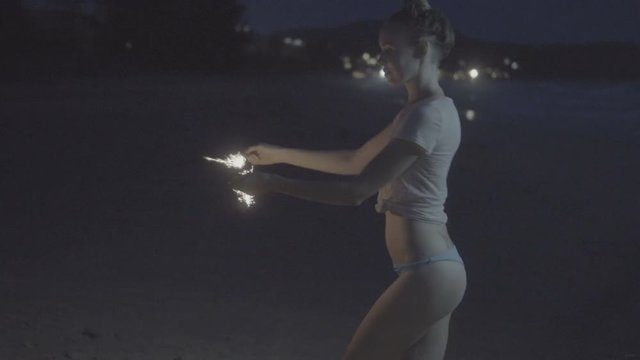 Side view of sensual woman wearing t-shirt and bikini bottom doing sparkler fireworks show on the beach at night