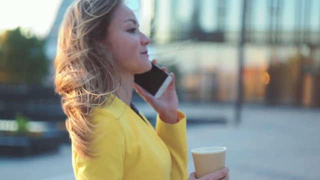 Camera moving around young beautiful business woman. Brunette girl talking by the phone and drinking coffee near office building in the big city. Cinematic style video.