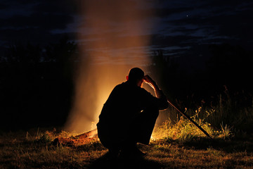 Silhouette of a man with a long stick sitting by the fire at nig