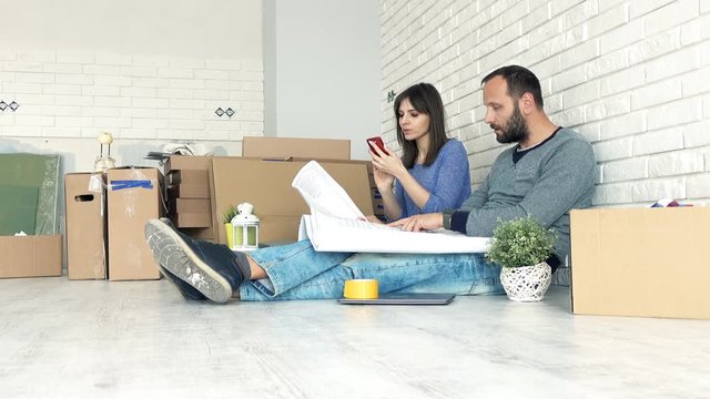 Young couple counting bills with smartphone and blueprints sitting on floor at their new home, 4K

