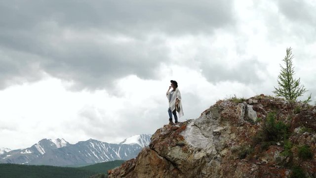 Young beautiful woman traveler wearing hat and poncho relaxing on the top of the hill with mountains and hills around