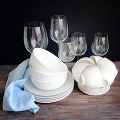 White dishware stacked on a wooden table against black background with transparent wineglasses
