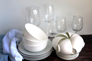 White dishware stacked on a wooden table against white background with transparent wineglasses. Special light.
