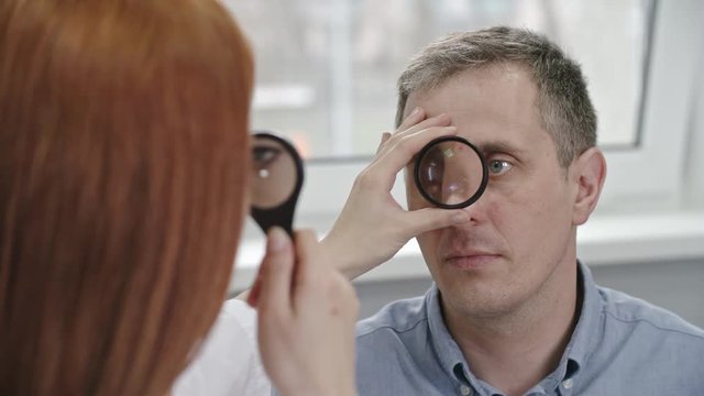 Closeup over the shoulder view of female optometrist checking eyes of mid adult patient through lens