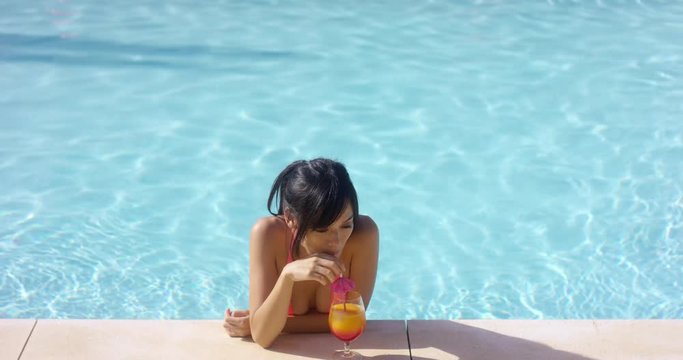 Contented beautiful single woman in black hair and bikini at pool edge with orange flavored drink and straw. Includes copy space.