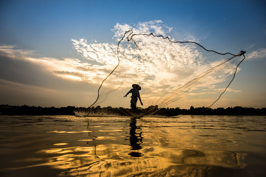 Asian fisherman on wooden boat casting a net for catching freshwater fish in nature river in the early morning before sunset