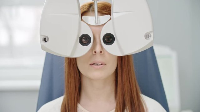 Closeup of young red-haired woman reading eye chart during distance vision test with modern automated phoropter 