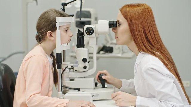 Side view of female pediatric optometrist examining eyes of little girl with modern biomicroscopic slit lamp device