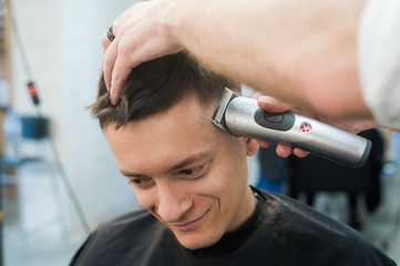 Professional styling. Close up side view of young man getting haircut by hairdresser with electric razor at barbershop