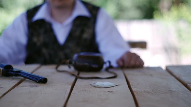 On the table lies a colt peacemaker, a flask and a deck of cards. In the background sits a man. Hand takes the card.