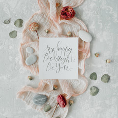 inspirational quote "be happy be bright be you" written in calligraphy style on paper with dry white tulips, eucalyptus petals and pink textile on concrete background. Flat lay, top view