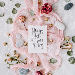 inspirational quote "follow your soul it knows the way" written in calligraphy style on paper with dry white tulips, eucalyptus petals and pink textile on concrete background. Flat lay, top view