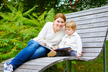Little boy and his mother sitting on bench in park and reading b