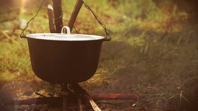 Camping scene: a pot with food being prepared hangs over the bonfire