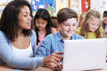 Teacher Helping Male Elementary Pupil In Computer Class