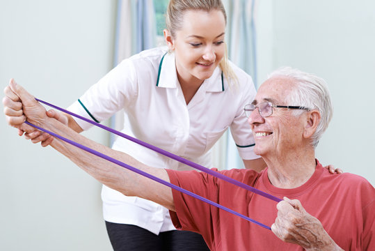 Physiotherapist Helping Senior Male To Use Resistance Band
