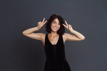 Cute young woman posing over grey background and make two peace funny gesture