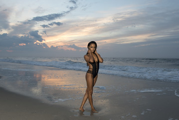Beautiful African American female model posing on beach in swimsuit at sunrise (sun rising behind her over ocean)