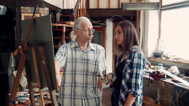 Senior Artist And young Woman painting on canvas using oil paint. He teaches young girl how to draw.