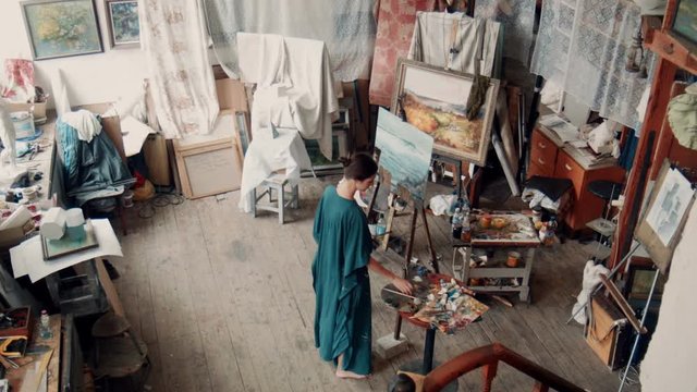 Aerial view of a female artist wearing a smock standing in her studio painting a canvas on an easel surrounded by other covered canvasses and art supplies.