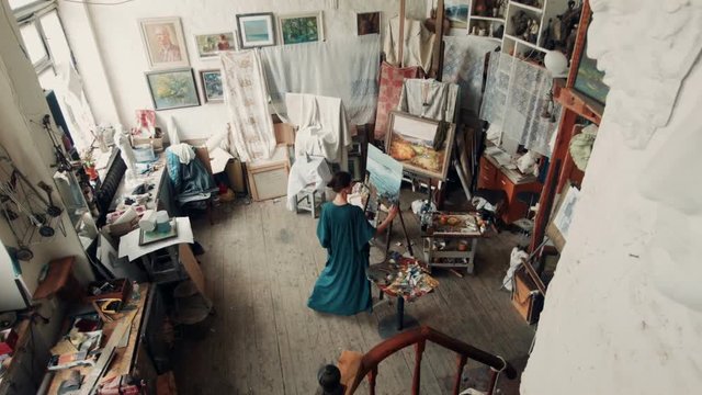 Wide view of a female artist wearing a smock standing in her studio painting a canvas on an easel surrounded by other covered canvasses and art supplies.