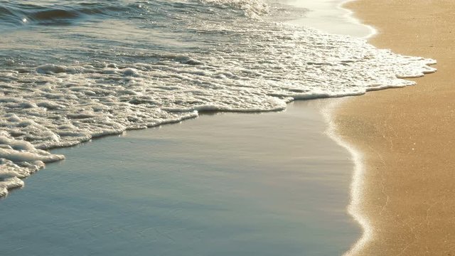 Close up of surf with soft sea waves rushing on sandy beach on sunset with sun reflection. Enjoying summer vacation on tropical country. Nature background