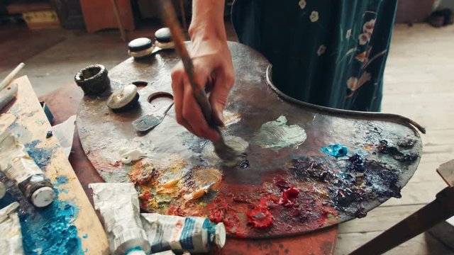 Woman artist mixing oil paint on a palette with a paintbrush from an array of different colored tubes surrounding it, close up of her hand and the art supplies