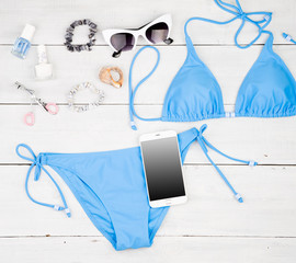 blue swimsuit, smart phone, cosmetics makeup, bijou and essentials on white wooden desk