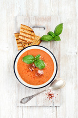 Cold tomato gazpacho soup in bowl with ice, hot pepper, basil and bread slices on wooden board over white wooden background, top view, vertical composition