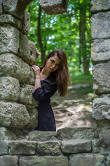 Young beautiful girl with long hair and a dark raincoat looks out the window of the old castle from which remained only ruins in Striysky Park in Lviv
