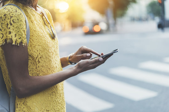 Close-up image of female hands using modern smart-phone while standing at a crosswalk,  side view of young hipster girl typing text message via cellphone outdoors, film effects, flare light