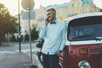 Happy smiling man talking on smartphone while standing near red classic bus, young hipster guy using cellphone while traveling around the world on a vintage bus