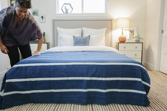 A woman smoothing a blue striped throw over a double bed in a bedroom.  