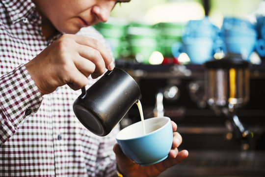 Specialist coffee shop. A man pouring hot milk into a cup of coffee to make a pattern on the top. 