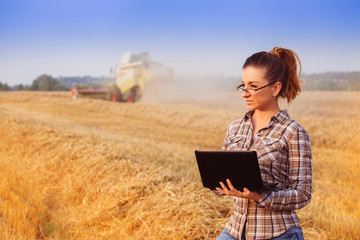 Young pretty farmer girl in glasses with hair tied in a ponytail to screw up one's eyes in wheat field while combine harvester working in background. Image released.