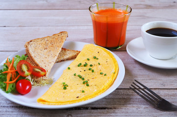 Omelette with toast, vegetable, carrot juice and coffee for breakfast on a wooden table.