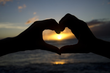 Women's hand on the left and man's hand on the right making a heart over the sunset.