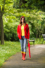 Portrait of a beautiful young woman in hooded red raincoat looking at camera and smiling while walking in the park on a rainy day.