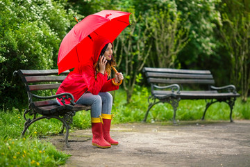 Beautiful smiling young woman in hooded red raincoat is holding red umbrella while sitting on a...