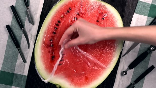 A half a red juicy ripe watermelon covering wrap is on the dark table. Melon is ready for desserts. Women's hand removes plastic wrap from water-melon. 4k