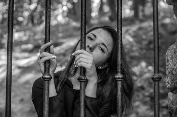 Young charming girl the teenager with long hair sitting behind bars in prison prisoner in a medieval jail with sad, pleading eyes of mercy