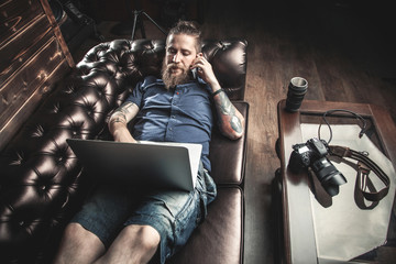 Male boss lying on the sofa in his office, working on laptop and talking on a cell phone. The concept of the creative process, skill and favorite pastime