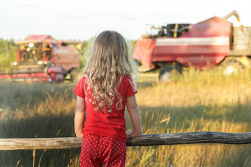 Little farm girl wearing red polka dot kids pans looking at field with working combine harvesters