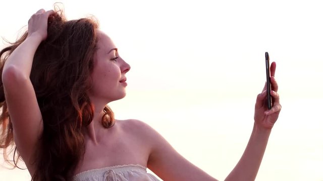 Selfies on the beach, beautiful girl does a mobile phone photo at sunset on the beach. Cheerful young woman taking selfies on exotic beach