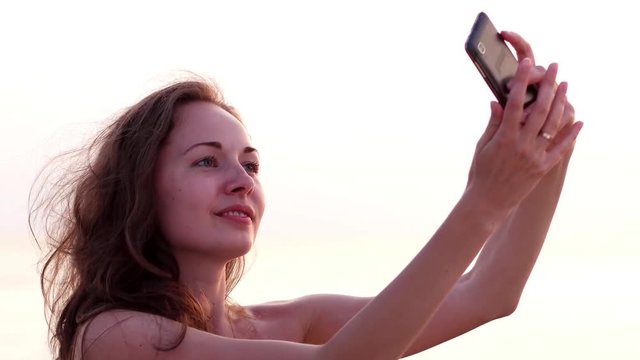 Beautiful, delicate, girl with long hair makes selfie, straightens hair, stroking her hair. girl in a white dress makes selfie phone on a background. selfie on the beach, photos to social networks,