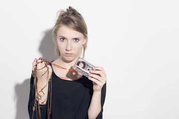 Young blonde woman damaging a cassette tape, looking to camera, over a white background.