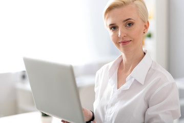 Woman looking at camera and holding laptop - obrazy, fototapety, plakaty