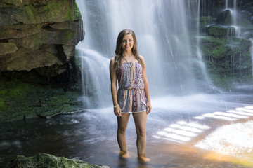 Beautiful young Caucasian woman stands in front of waterfall in a shallow pool wearing brightly colored romper - long exposure - art or fashion